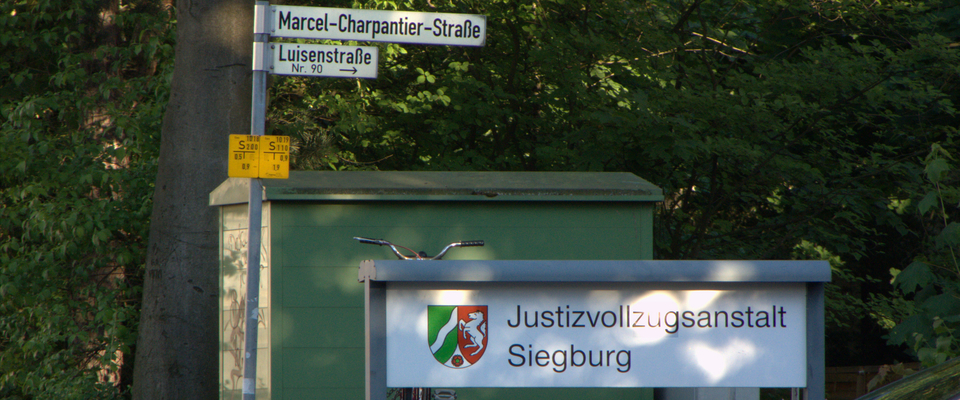 An der Einfahrt zur JVA Siegburg steht ein Schild mit dem Hinweis "Justizvollzugsanstalt Siegburg". Das Straßenschild weist auf die Luisenstr. 90 und die Marcel-Charpantier-Straße hin.