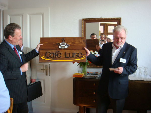 Der Anstaltsleiter der Justizvollzugsanstalt Siegburg Herr Wolfgang Klein und der Anstaltspfarrer Herr Werner Kaser überbringen ein in der Arbeitstherapie der Justizvollzugsanstalt Siegburg  hergestelltes Holzschild für das Café Luise.