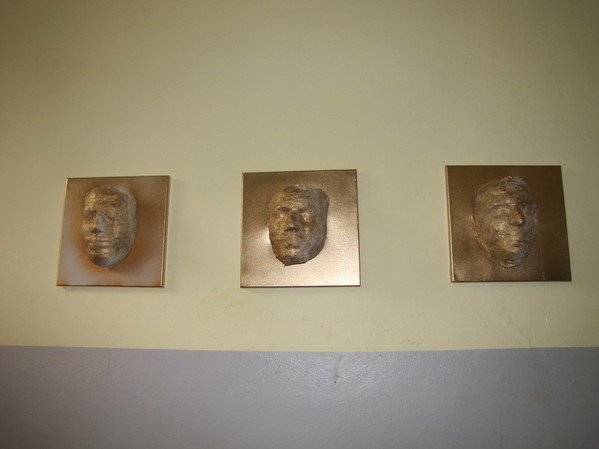 Drei kupferfarbene Masken an der Zellenwand.