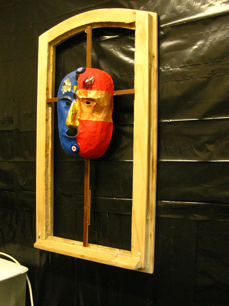 Eine Maske im Holzrahmen mit zwei Gesichtsseiten einer Blauen und einer Roten. Mit einer goldenen Nase auf schwarzem Hintergrund.